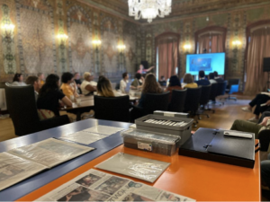 Feature image from the Community Archiving Workshop in Istanbul, Turkey, 14 September 2023. Collection materials are in foreground in an opulent room with people gathered around a long table during a presentation in background.