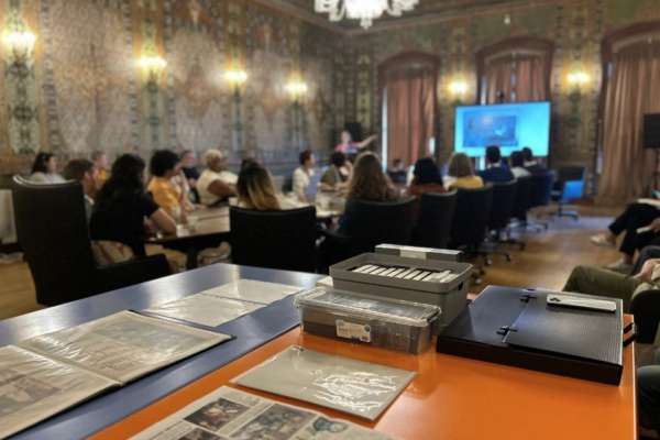 Feature image from the Community Archiving Workshop in Istanbul, Turkey, 14 September 2023. Collection materials are in foreground in an opulent room with people gathered around a long table during a presentation in background.