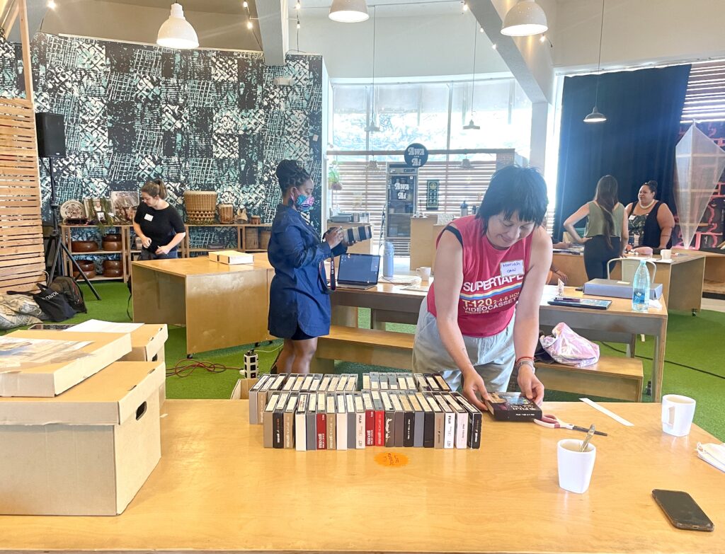 Image from 2023 workshop in Honolulu. Setting up the space, organizing a collection of video tapes on a table. People in the background talk.