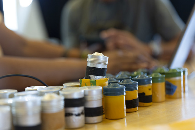 Cans of 35mm still image film lines up on a table in the foreground while hands type on a computer in the background.